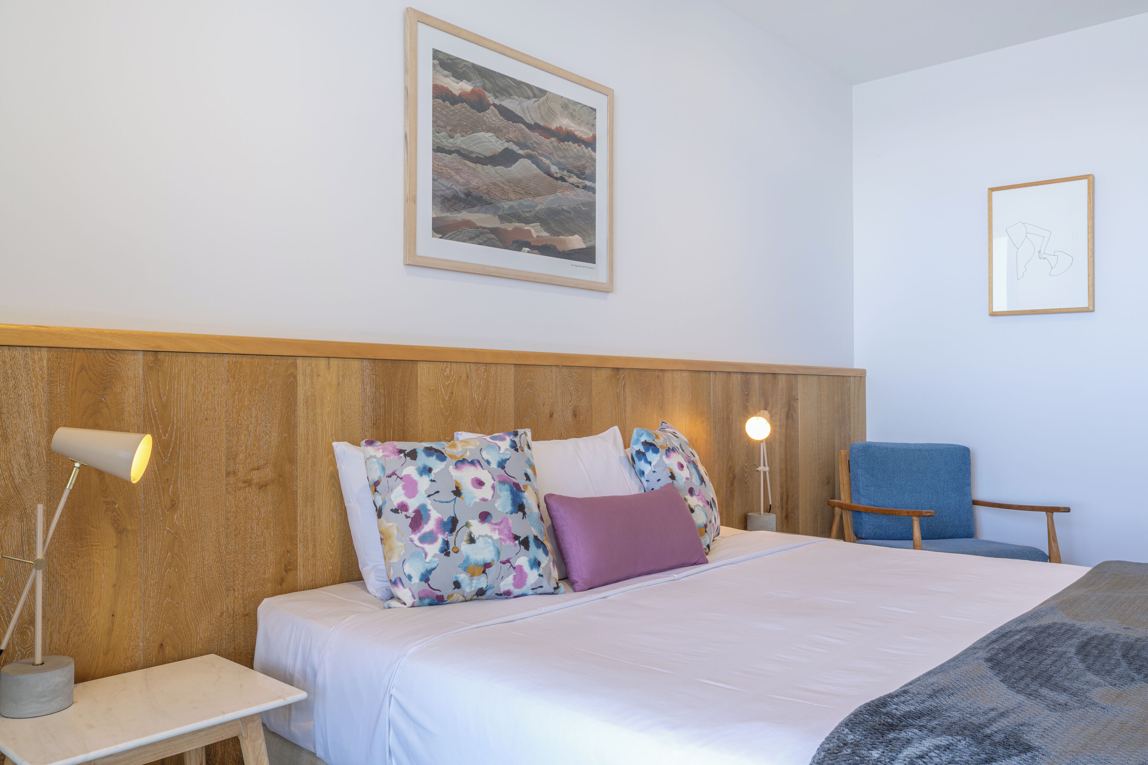 A neatly arranged hotel room with a bed, a chair, a desk, and a lamp.