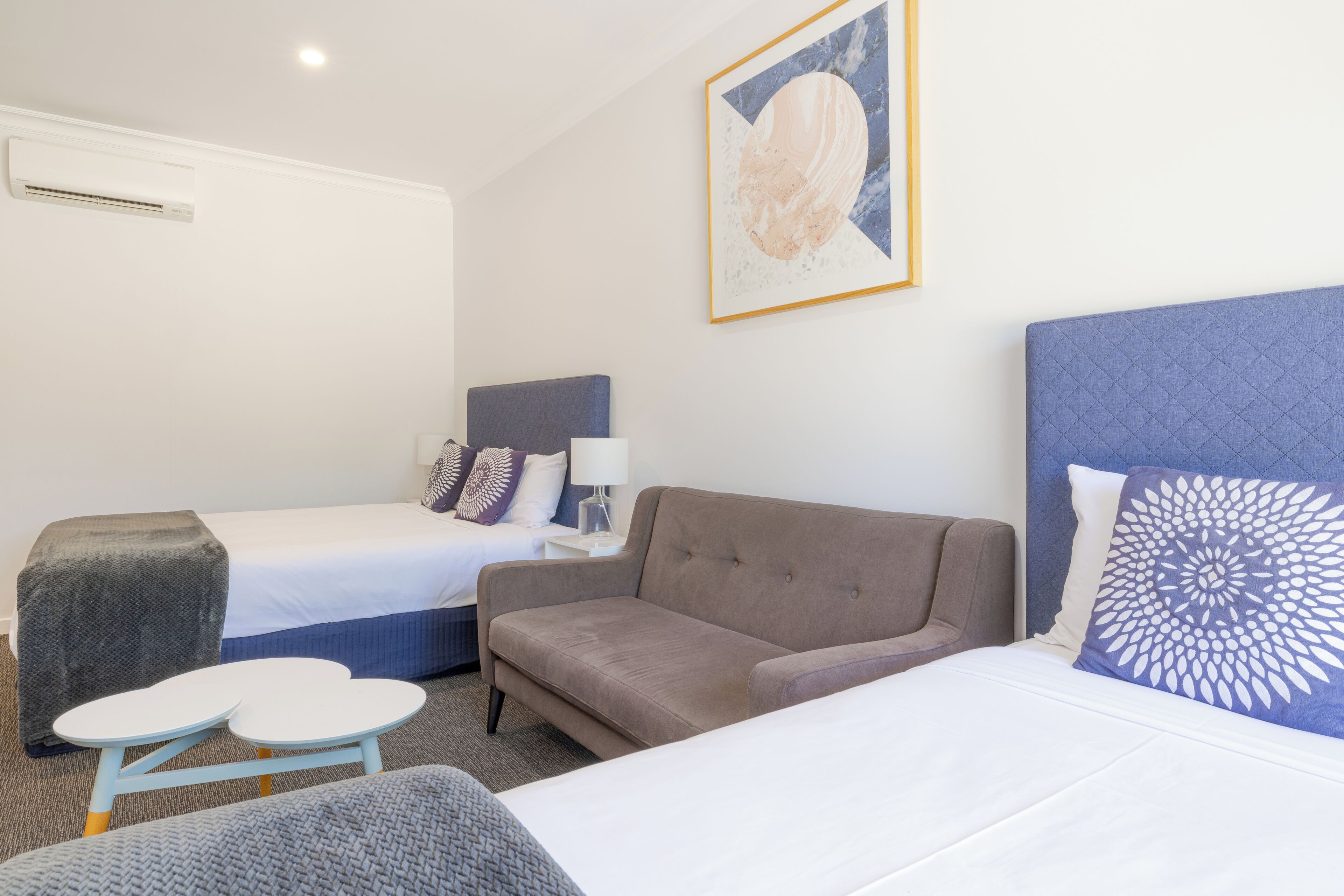 A hotel room with two beds, a sofa, a small table, and a wall-mounted air conditioner.