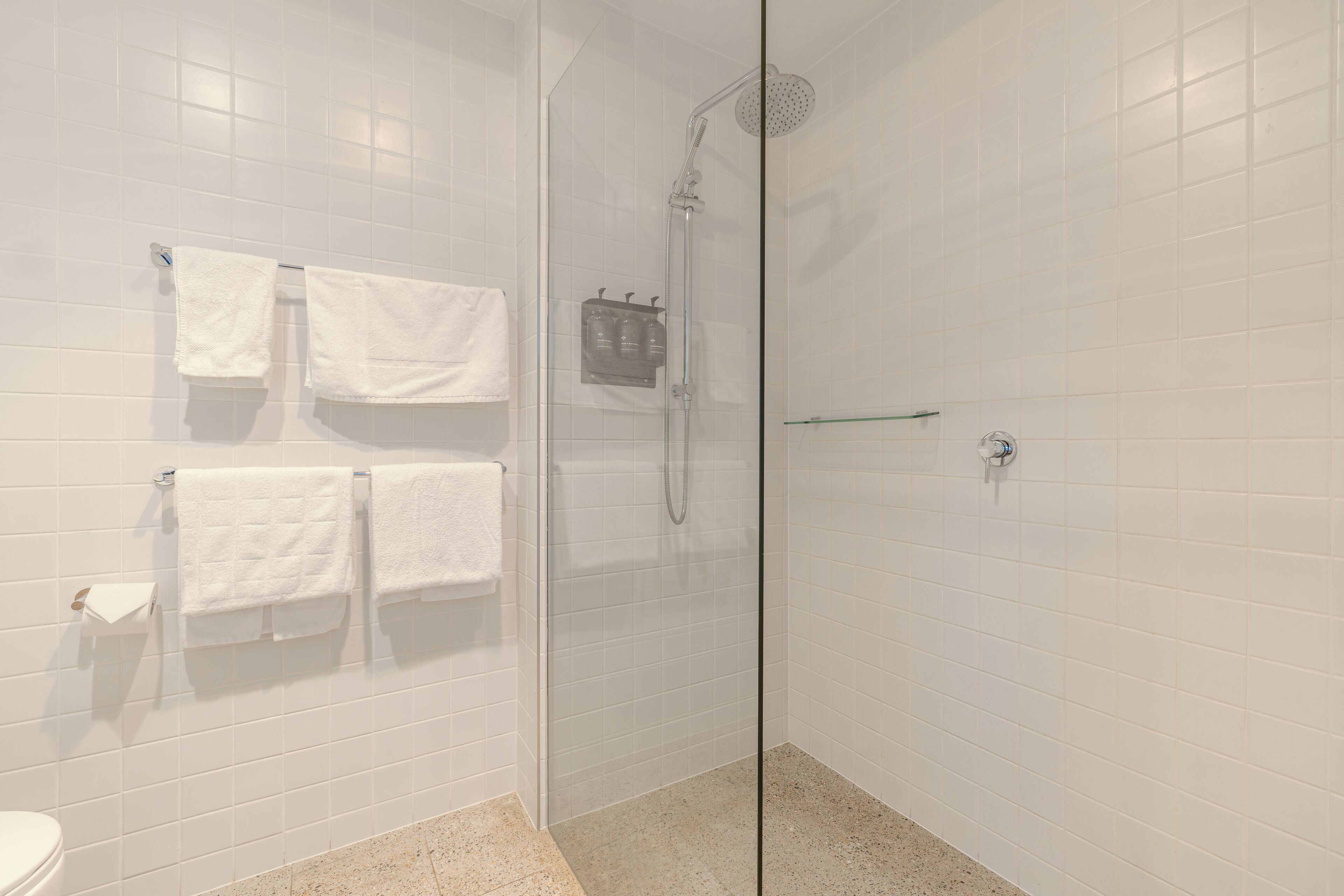 Un baño moderno con paredes de azulejos blancos, una cabina de ducha de vidrio y varias toallas blancas colgadas en un perchero.