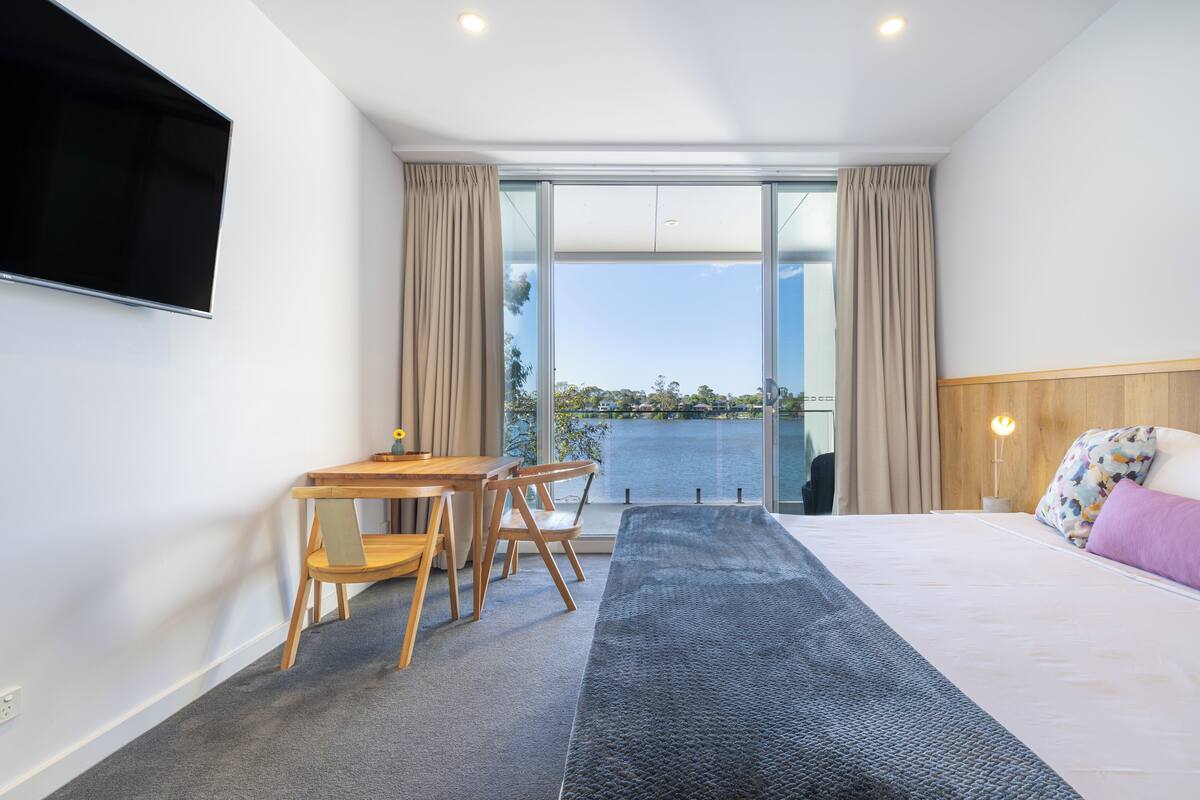 Una habitación de hotel moderna con una cama grande, un escritorio con sillas, un televisor de pantalla plana y vista al agua.