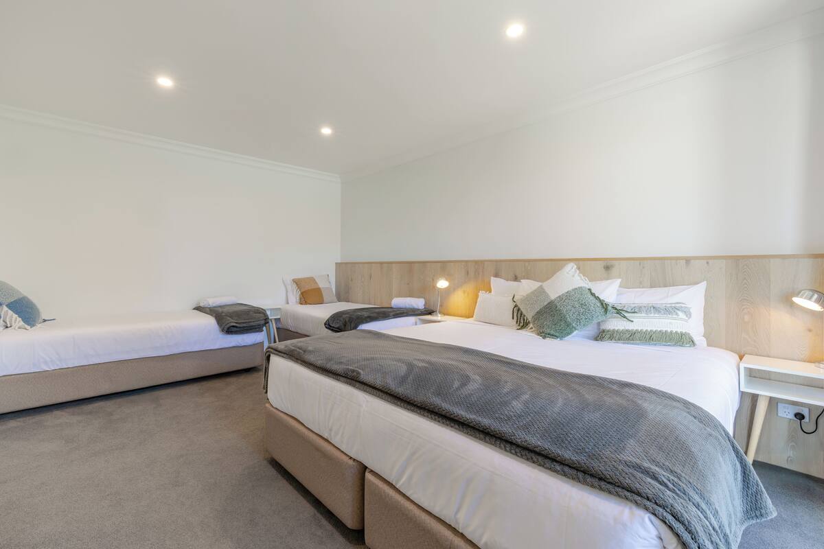 Habitación de hotel con dos camas individuales, una cama más grande, cabecera de madera, mesitas de noche con lámparas y suelo cubierto de alfombra.