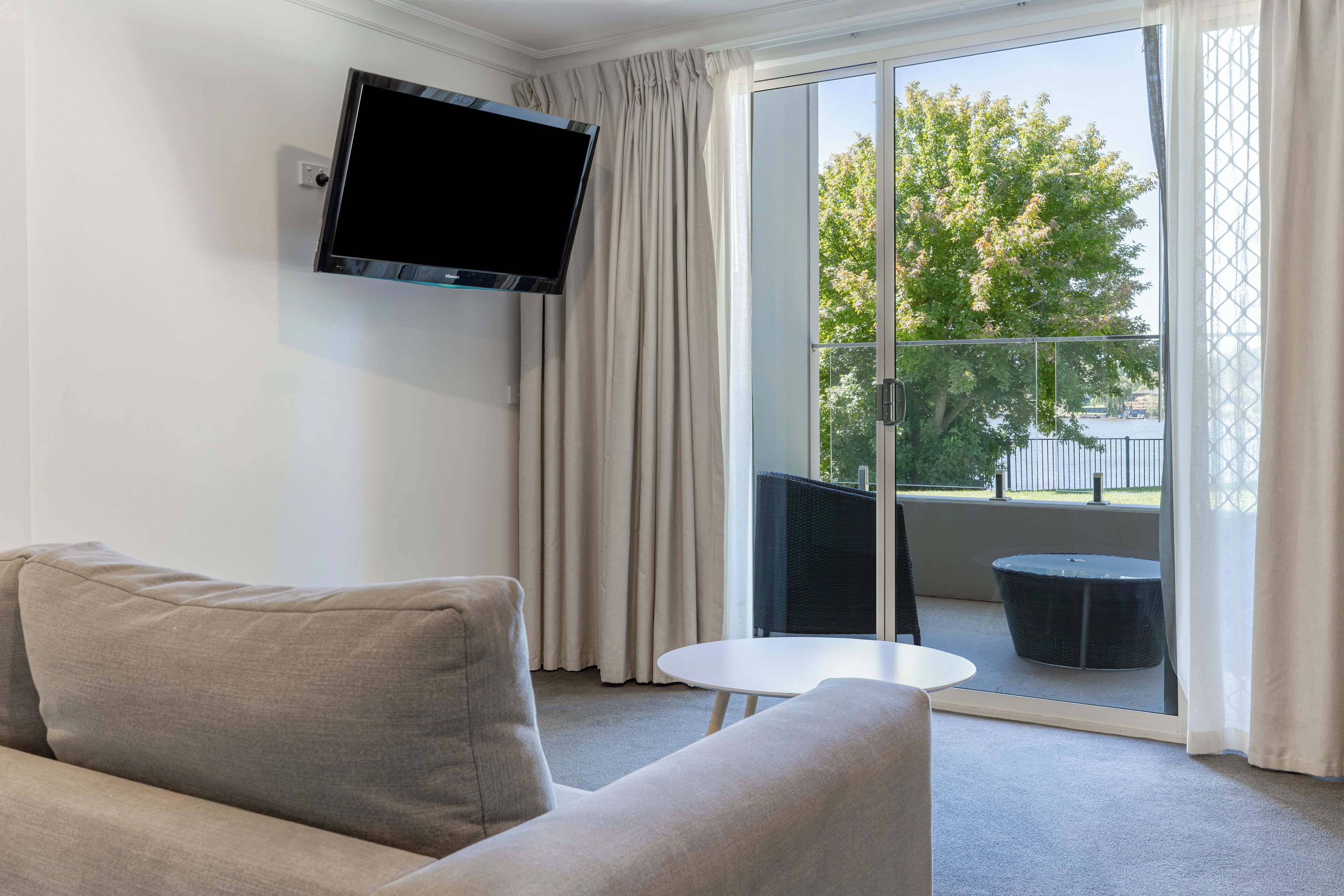 A modern living room with a sofa, a flat-screen TV mounted on the wall, and a balcony with a table and chairs.