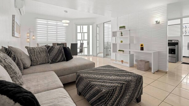 Ein modernes Wohnzimmer mit einer grauen Couch, einem gemusterten Hocker und einer weißen Regaleinheit mit Dekorationselementen.