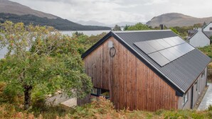 A wooden cabin with a sloped roof, a small window, and a door, surrounded by trees and a lake. - Tigh Na Allt by Interhome (Mid Skye)
