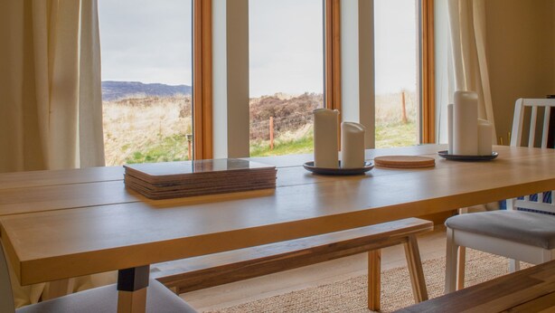 A room with a large wooden table, two white candles, and a view of a grassy landscape through three windows. - Vacation home Solace by Interhome (North Skye)