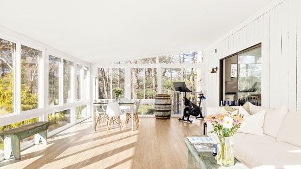 Una habitación luminosa e inundada de sol, con ventanas amplias, piso de madera, un sofá blanco, una mesa de comedor con sillas y una mesa de centro con un jarrón de flores.