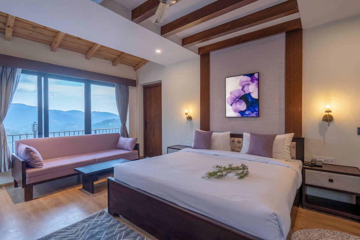 A modern hotel room with a large bed, a sofa, a view of mountains, and a painting on the wall.