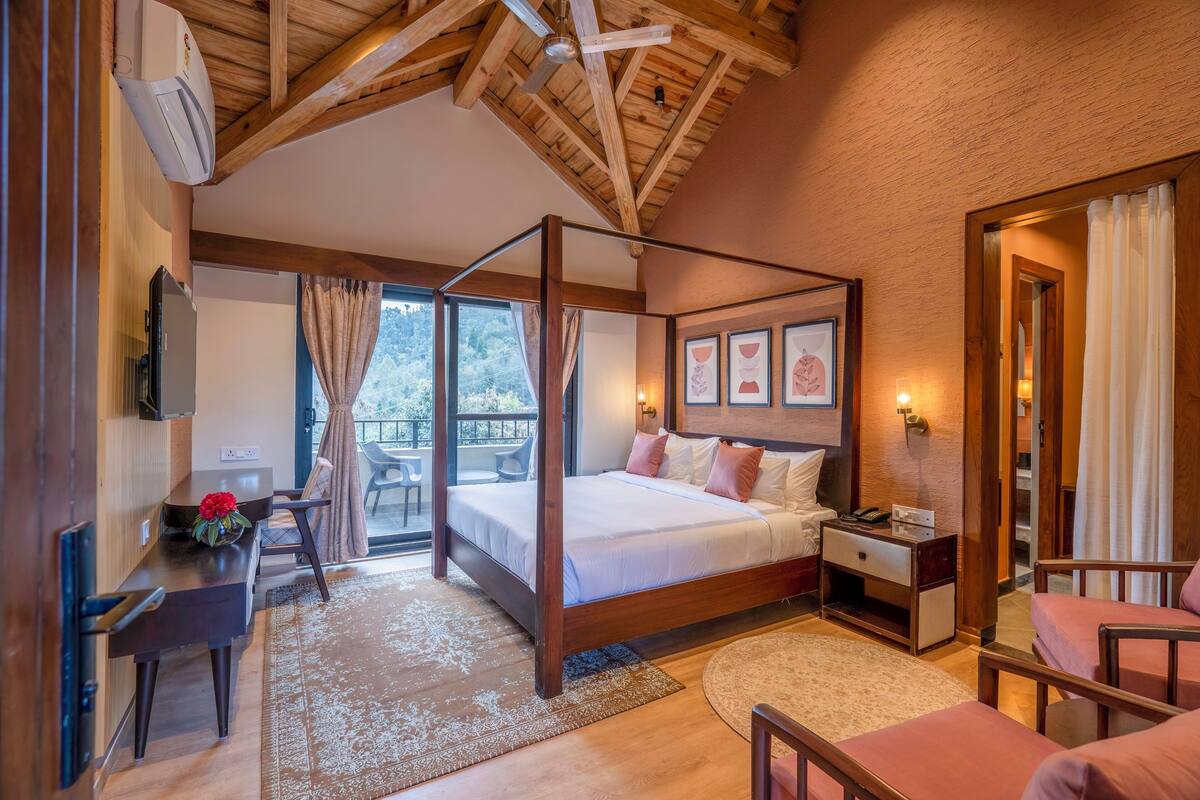A four-poster bed with a wooden frame, a desk with a chair, a small table with a vase of flowers, and a balcony with a chair and table.