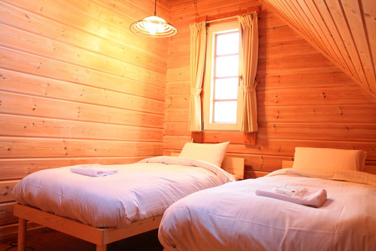 Una habitación de cabaña de madera con dos camas, una ventana con cortinas y una lámpara colgante.
