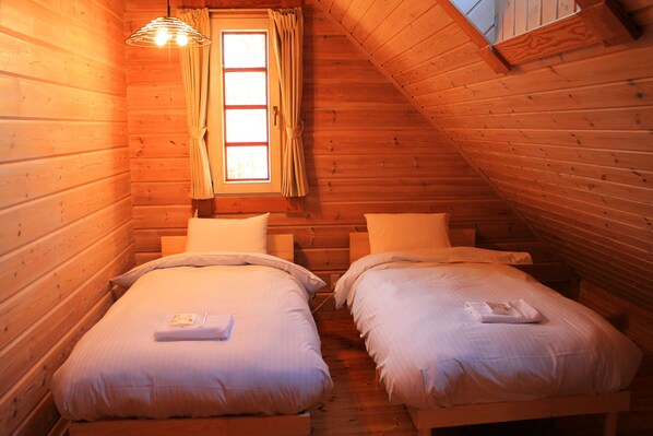 A cozy attic bedroom with two beds, wooden walls, a window with curtains, and a hanging light fixture.