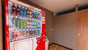 Ein Getränkeautomat mit verschiedenen Getränken, darunter Coca-Cola und Fanta, in einem Raum mit Holzboden und einer Tür.