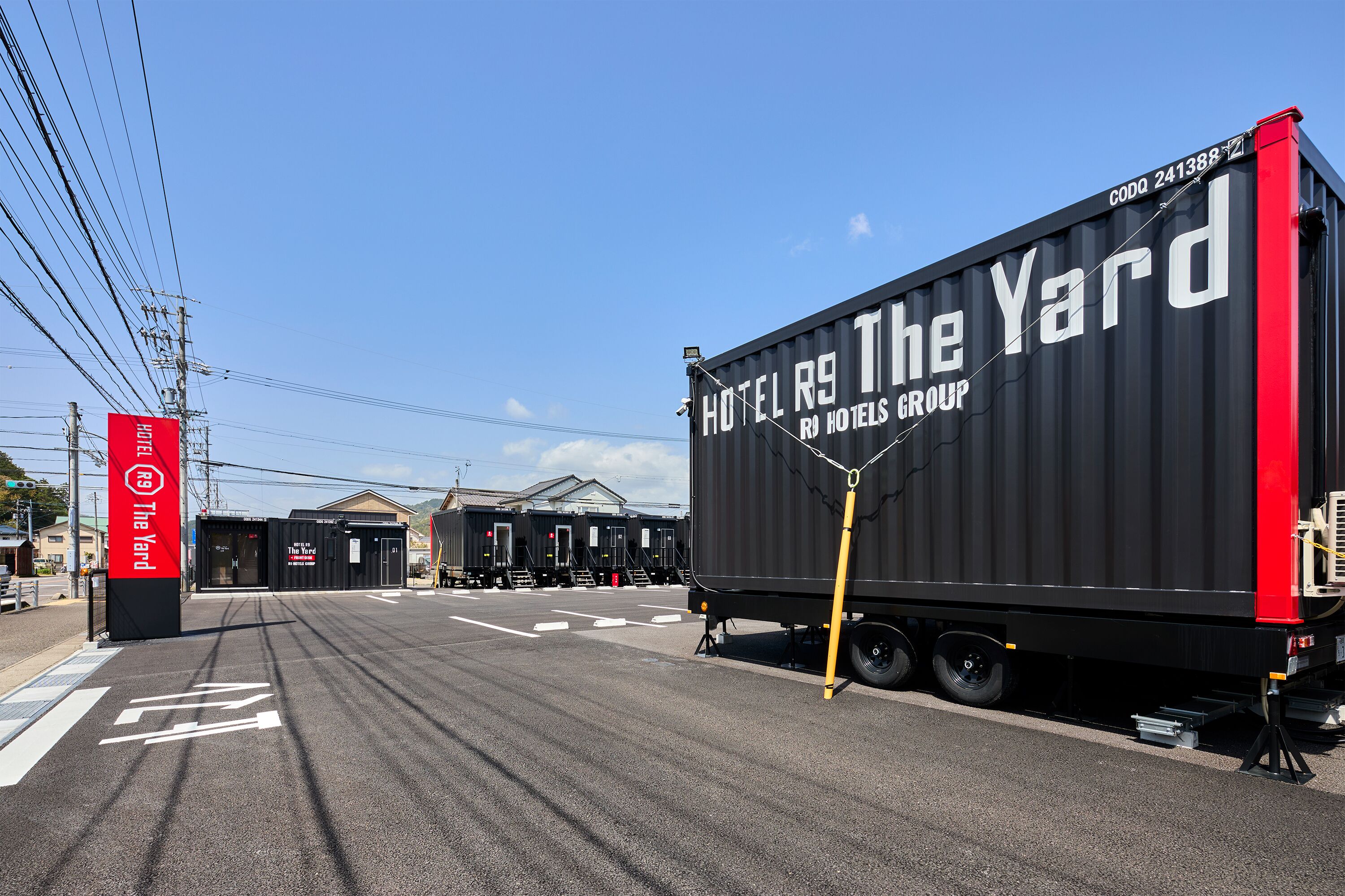 Un conteneur noir sur lequel on peut lire « HOTEL R9 The Yard » et « HOTEL GROUP », garé dans une rue sous un ciel dégagé.