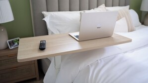 A bed with a laptop on a wooden bedside table, a remote control, and a nightstand with a lamp and a picture frame.