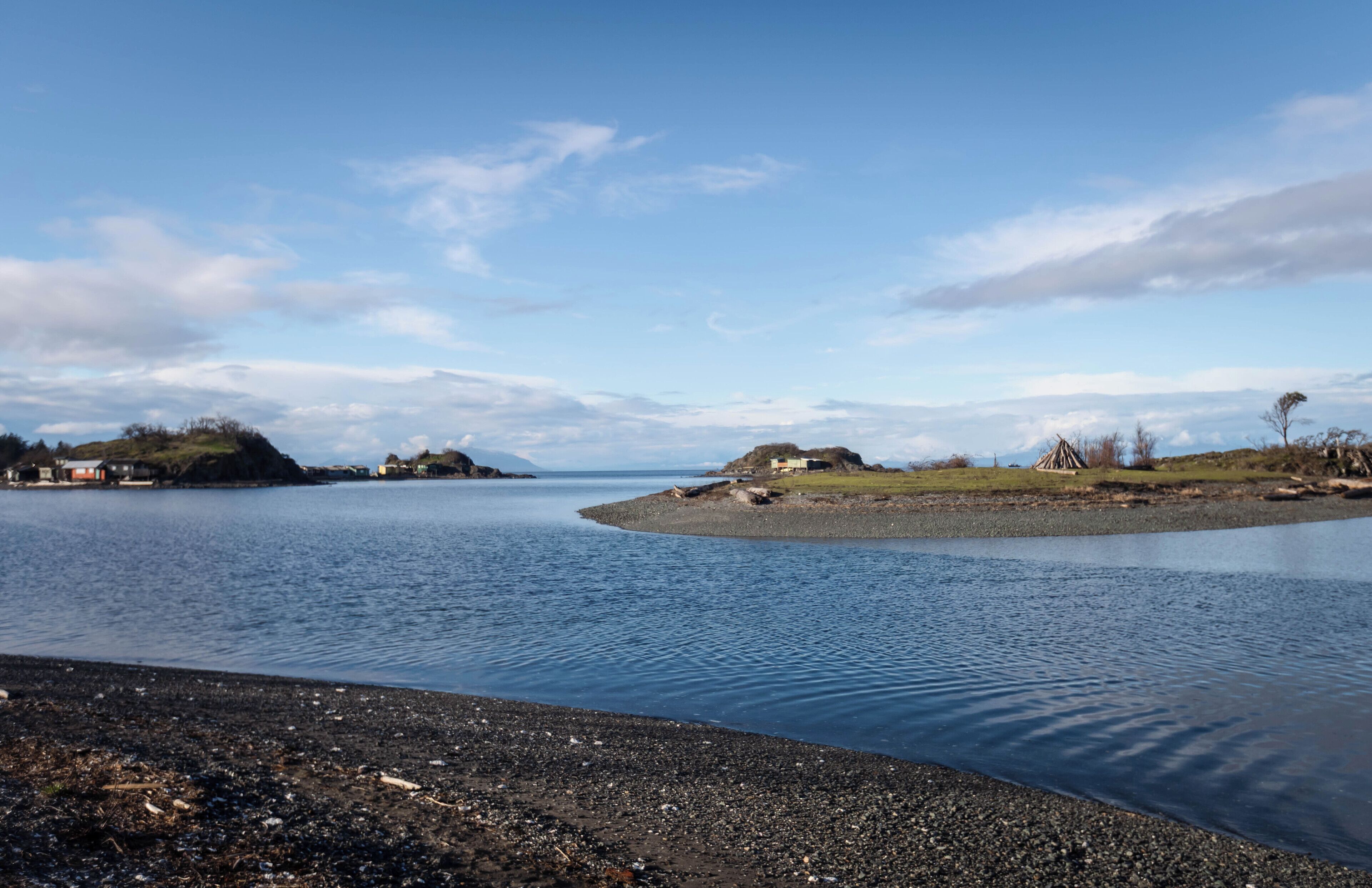 A calm lake with a pebbly shore and small buildings on the opposite bank.