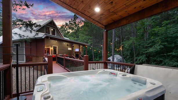 A hot tub on a wooden deck with a view of a cabin and forest. - Luxury Romantic Cabin - KingBed ~ Strip Lounge (Broken Bow)