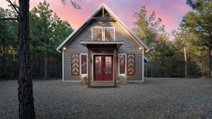 A wooden cabin with a red door, set in a forested area with gravel ground. - Luxury Romantic Cabin - KingBed ~ Strip Lounge (Broken Bow)
