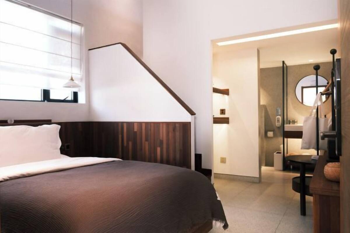 A modern hotel room with a wooden headboard, a large bed, a window with blinds, and a bathroom visible through an open door.