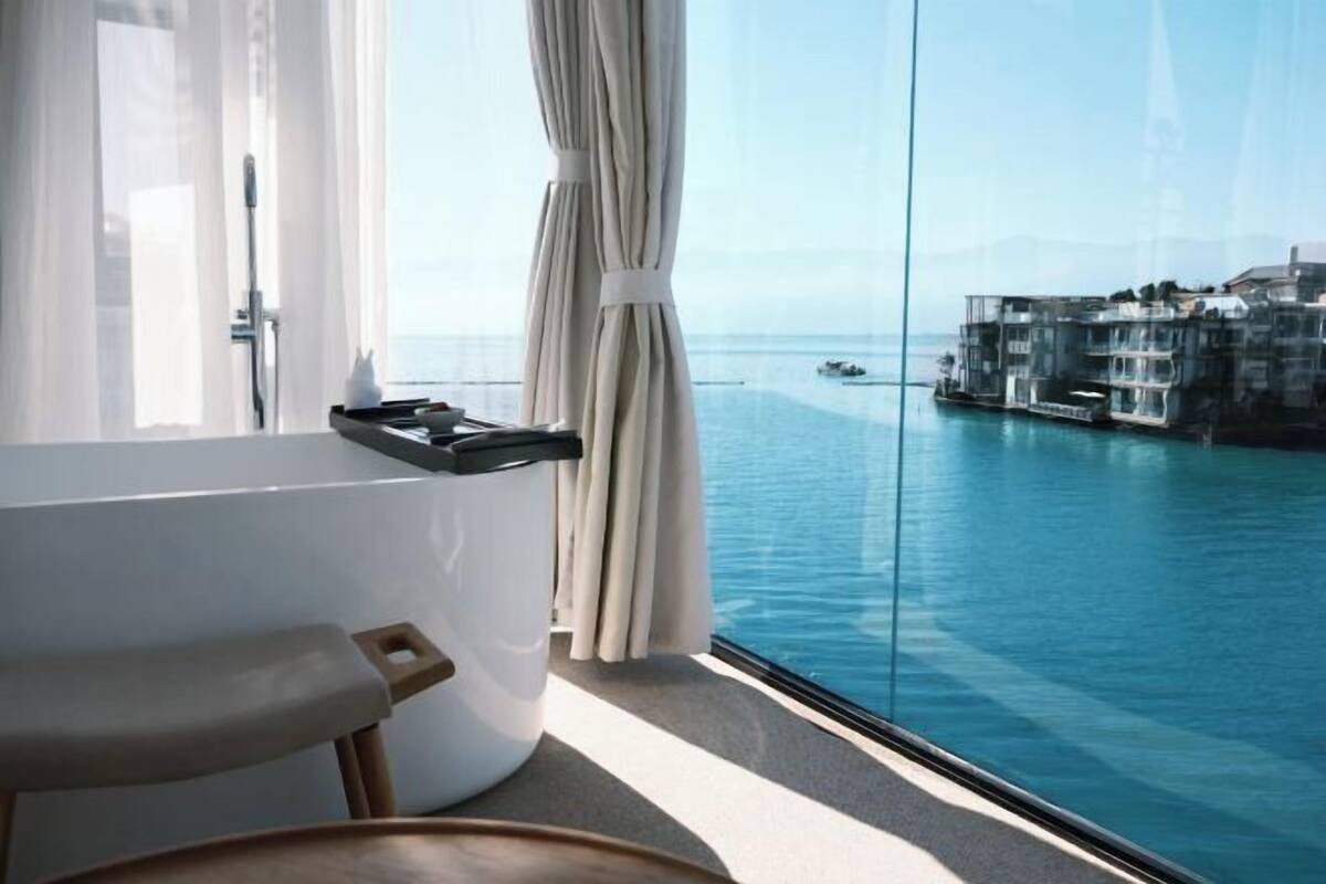 A modern bathroom with a bathtub, a shower, and a view of a coastal town through a large window.