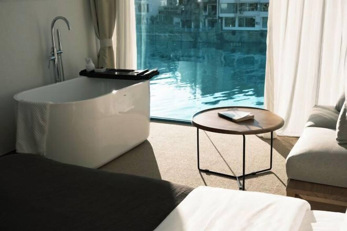 A modern bathroom with a freestanding bathtub, a round table, and a view of a body of water through large windows.