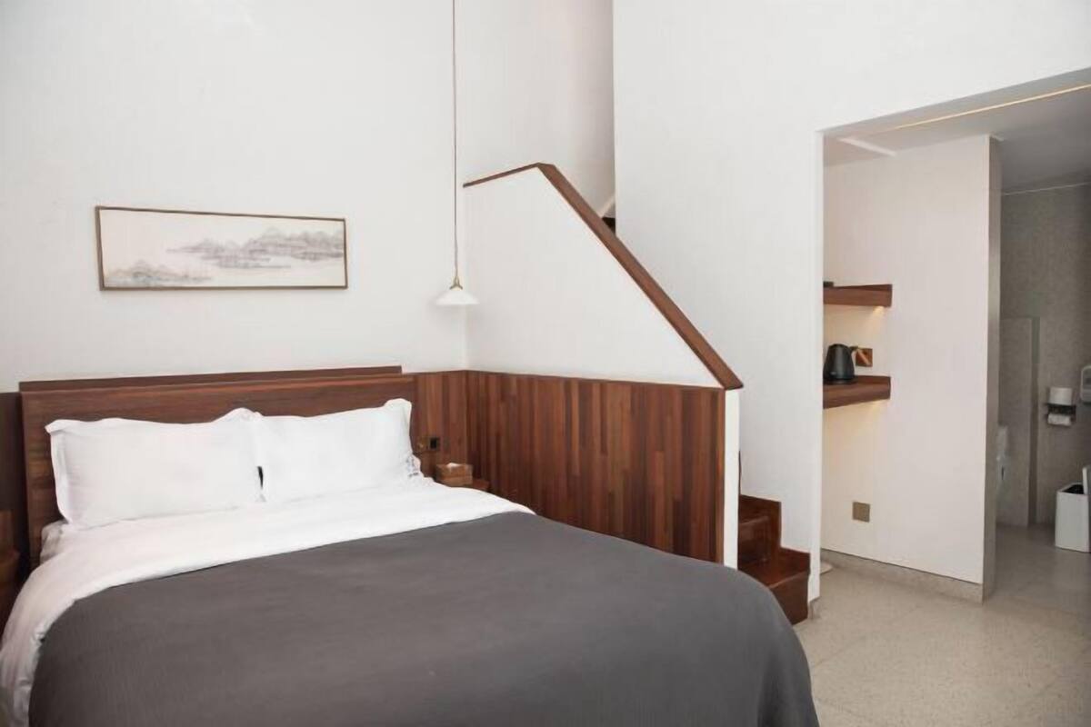 A bedroom with a wooden headboard, a bed with white bedding, a nightstand, a wall-mounted lamp, and a staircase leading to a kitchen area.
