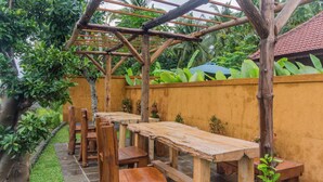 An outdoor wooden pergola with tables and chairs, surrounded by a yellow wall and lush greenery. - Green Zone Villas Ubud (Ubud)