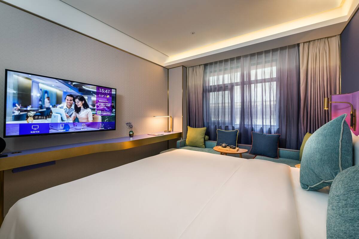 Habitación de hotel con una cama grande, televisor montado en la pared, un sofá y una ventana con cortinas.