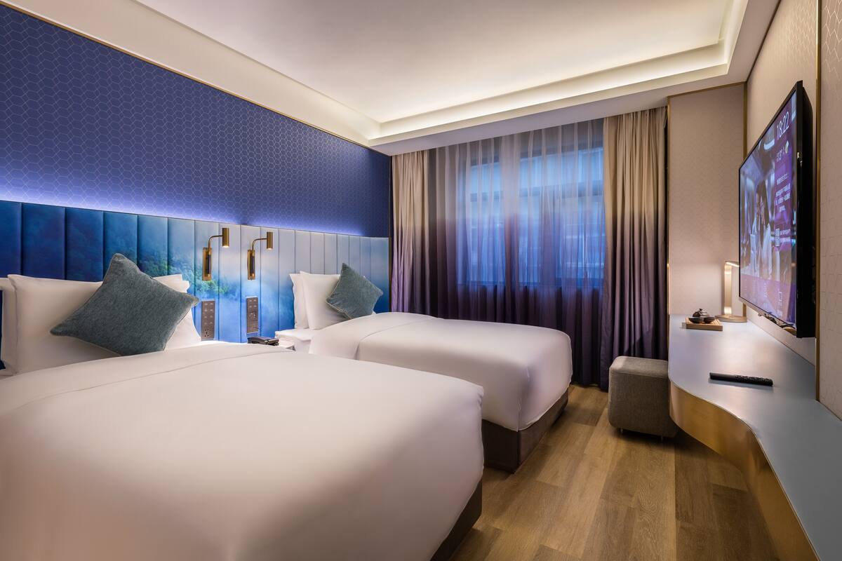 Habitación de hotel con dos camas, un televisor, una mesita pequeña y una ventana con cortinas.