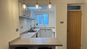 Eine moderne Küche mit einer zentralen Kochinsel, zwei schwarzen Stühlen, Pendelleuchten und Blick auf die Stadt durch ein Fenster.