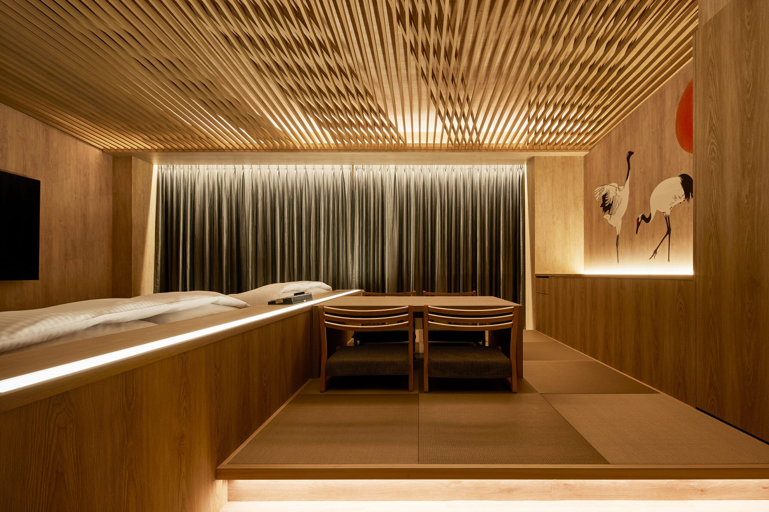 A room with a wooden ceiling, a bed with a headboard, a table with chairs, and a wall with a painting.