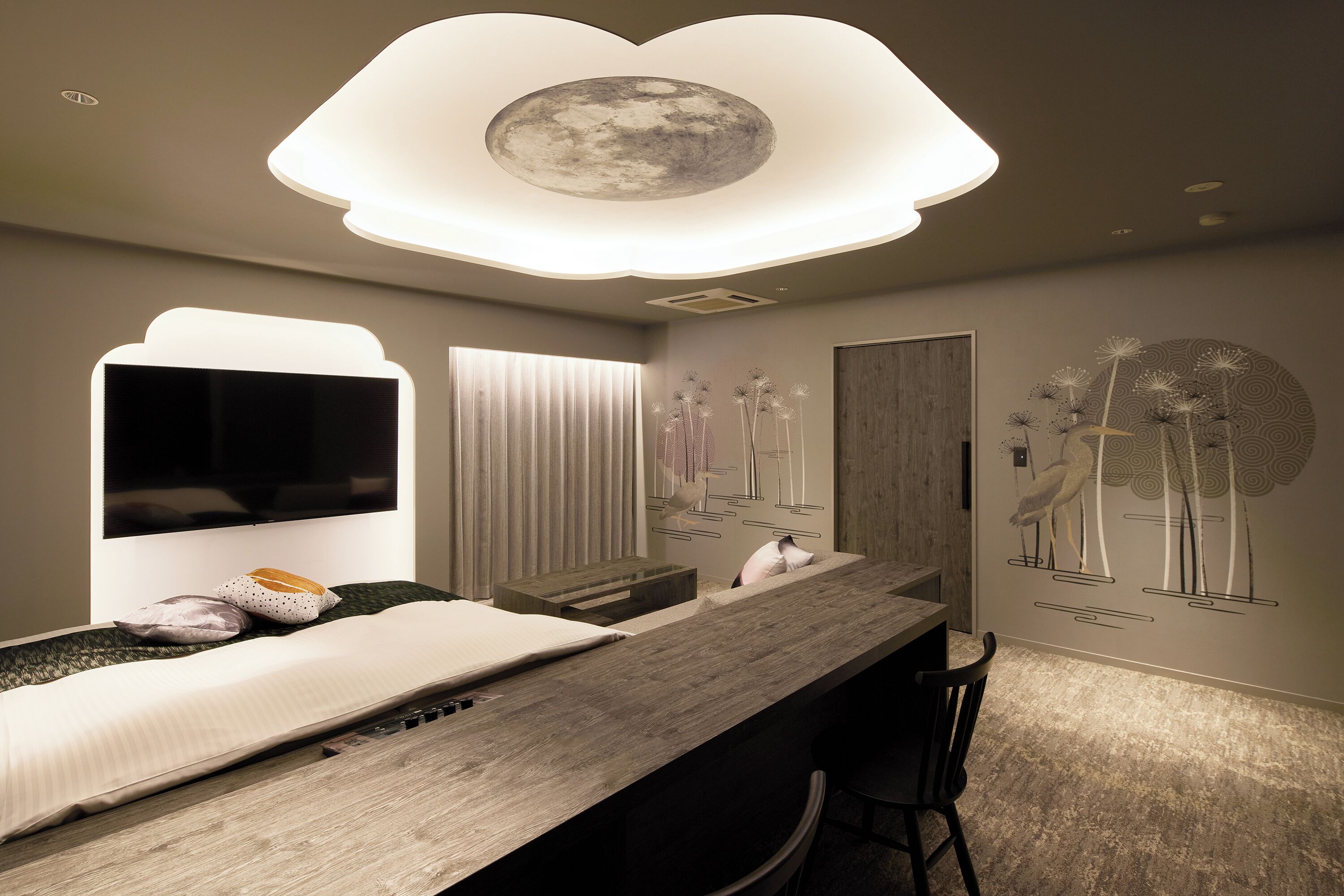A modern bedroom with a wooden desk, a flat-screen TV, a bed with pillows, and a ceiling light with a moon design.