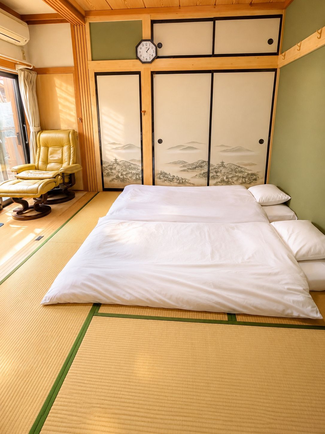 A traditional Japanese-style room with tatami flooring, a bed with white bedding, a yellow chair, and a wall with a clock and a landscape painting.