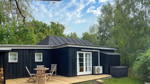 A dark wooden cabin with a deck, a large window, and an air conditioning unit. - Cozy home in Vejby with WiFi (Vejby)