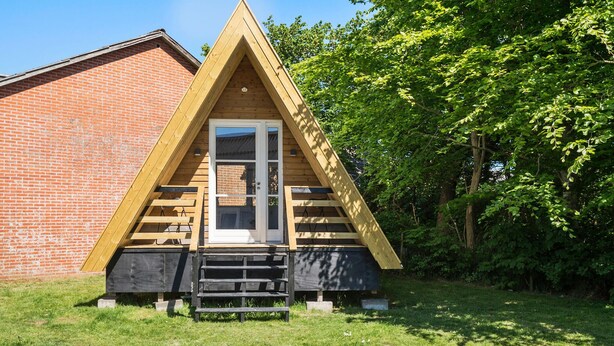A triangular wooden cabin with a brick wall adjacent to it, set in a grassy area with trees in the background. - Stunning home in Vemb with WiFi (Vemb)