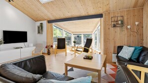 A modern living room with a large TV, comfortable seating, a wooden ceiling, and large windows.