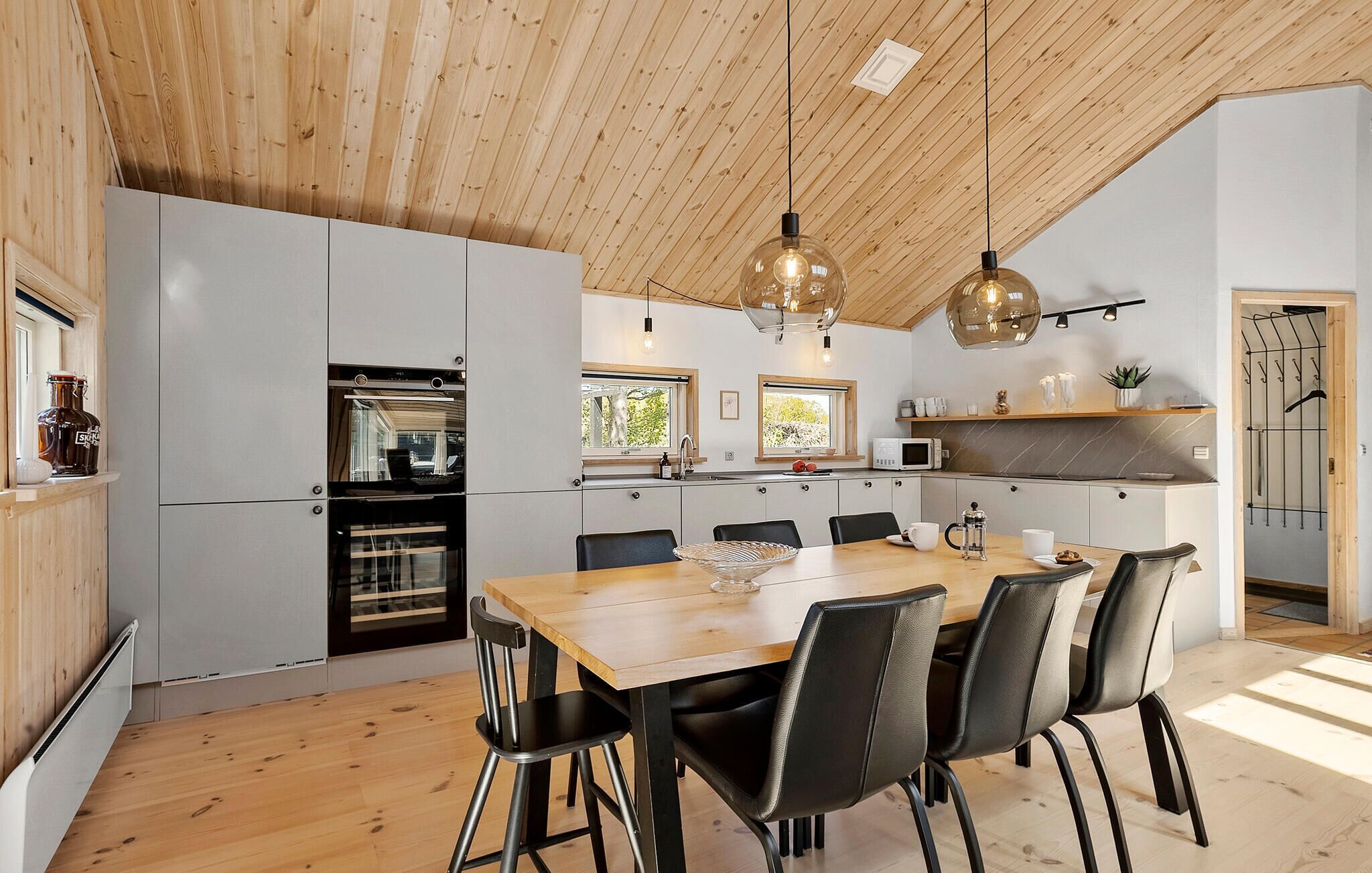 Una cocina moderna con techo de madera, un comedor con mesa de madera y sillas negras, y vistas al exterior a través de las ventanas.