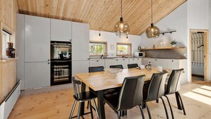 A modern kitchen with a wooden ceiling, a dining area with a wooden table and black chairs, and a view of the outdoors through the windows.