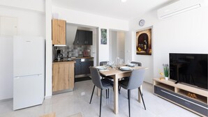 Een moderne keuken met een VIVAX koelkast, een houten kast en een betegelde spatwand. Er is een eethoek met een tafel gedekt voor vier personen, stoelen en een televisie op een houten standaard.