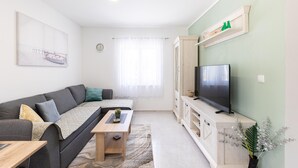 Ein modernes Wohnzimmer mit einem grauen Sofa, einem Holztisch, einem Flachbildfernseher auf einem Holzgestell und einem großen Fenster mit durchscheinenden Vorhängen.