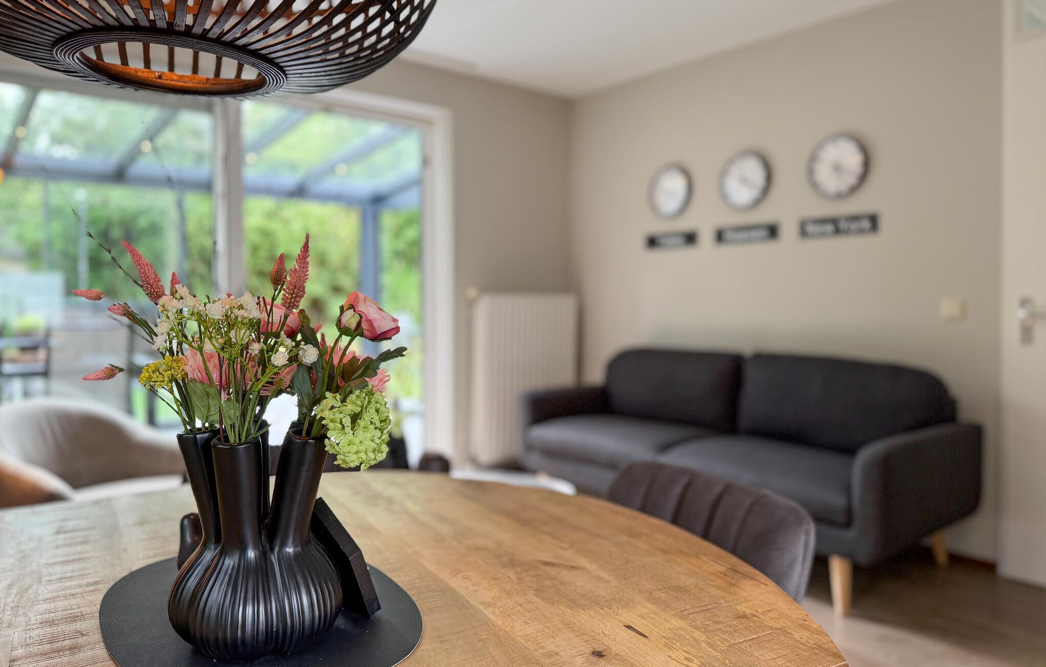 Een eetgedeelte met een houten tafel, drie zwarte vazen met bloemen, een moderne hanglamp en een grijze bank op de achtergrond.