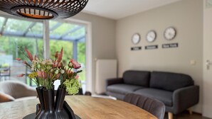 Un coin repas avec une table en bois, trois vases noirs ornés de fleurs, un luminaire suspendu moderne et, au fond, un canapé gris.