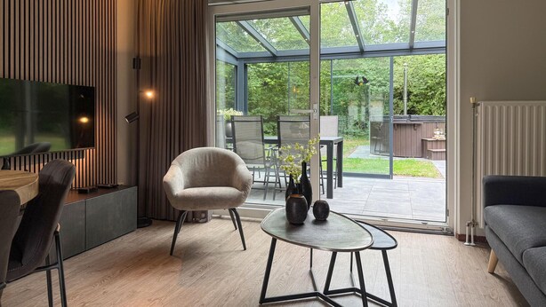 Un salon moderne doté d’un grand téléviseur, de sièges confortables, d’une porte vitrée donnant sur un jardin et d’un coin repas visible à travers la vitre.