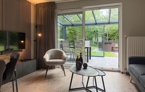A modern living room with a large TV, comfortable seating, a glass door leading to a garden, and a dining area visible through the glass. - 3 bedroom stunning home in Hoeven (Hoeven)