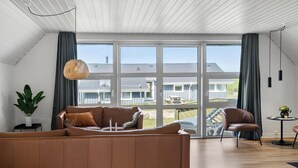 A spacious living room with a large window, a brown leather sofa, a dining table with chairs, and a hanging light fixture.