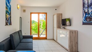 En moderne stue med en grå sofa, et fladskærms-tv på et træskab, og en skydedør med glas der fører ud til en balkon med grønt.