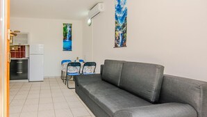 En stue med en grå sofa, et spisebord med stole, et køkken med et køleskab og to billeder hængt på væggen.