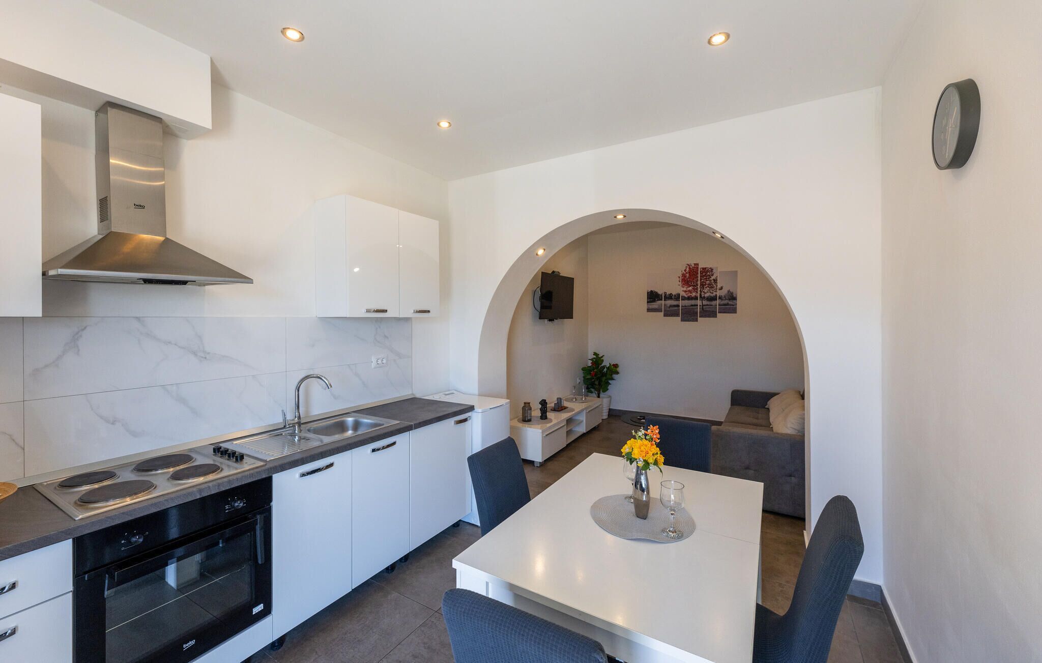 A modern kitchen with white cabinets, a built-in oven, and a stove. There is a dining area with a table and chairs, and a living space with a sofa and a TV.