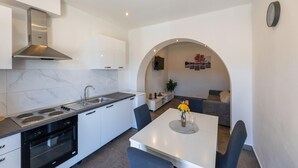 A modern kitchen with white cabinets, a built-in oven, and a stove. There is a dining area with a table and chairs, and a living space with a sofa and a TV. - Lovely apartment in Ploce with WiFi (Ploce)