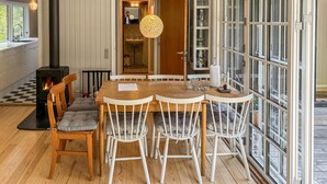 Un coin repas avec une table en bois et des chaises blanches, un poêle à bois noir et de grandes fenêtres donnant sur l’extérieur.