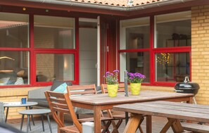 Ein Terrassenbereich im Freien mit Holztischen und -stühlen, großen Fenstern mit roten Rahmen und Kübelpflanzen mit violetten Blüten.