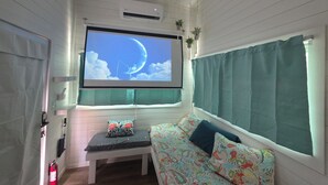 A cozy room with a large flat-screen TV mounted on a wall, a bench, and a sofa with patterned cushions. - Blackwater Bay beach tiny homes (Bolivar Peninsula)